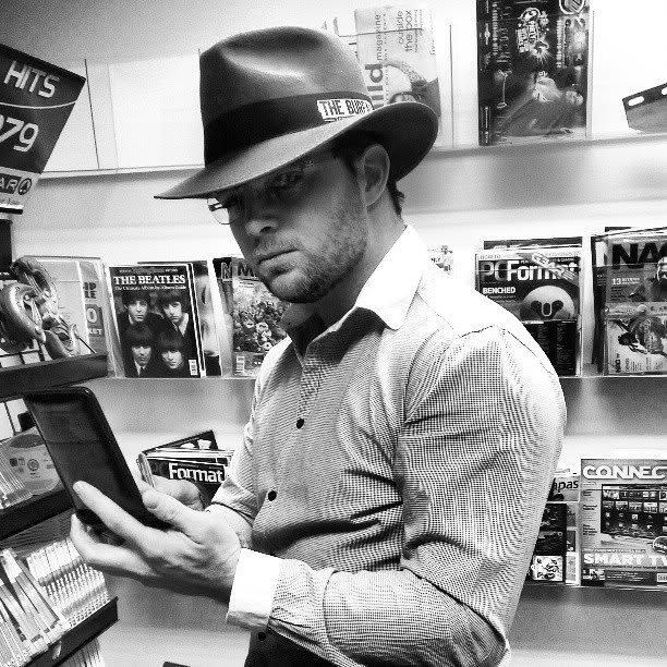 Devon in a fedora reading a tablet in front of a magazine rack, in black and white